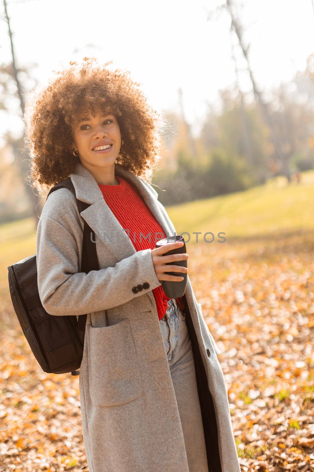 A joyful woman with curly hair strolls through a sunny park adorned with colorful autumn leaves, holding a warm cup of coffee while wearing a stylish outfit.