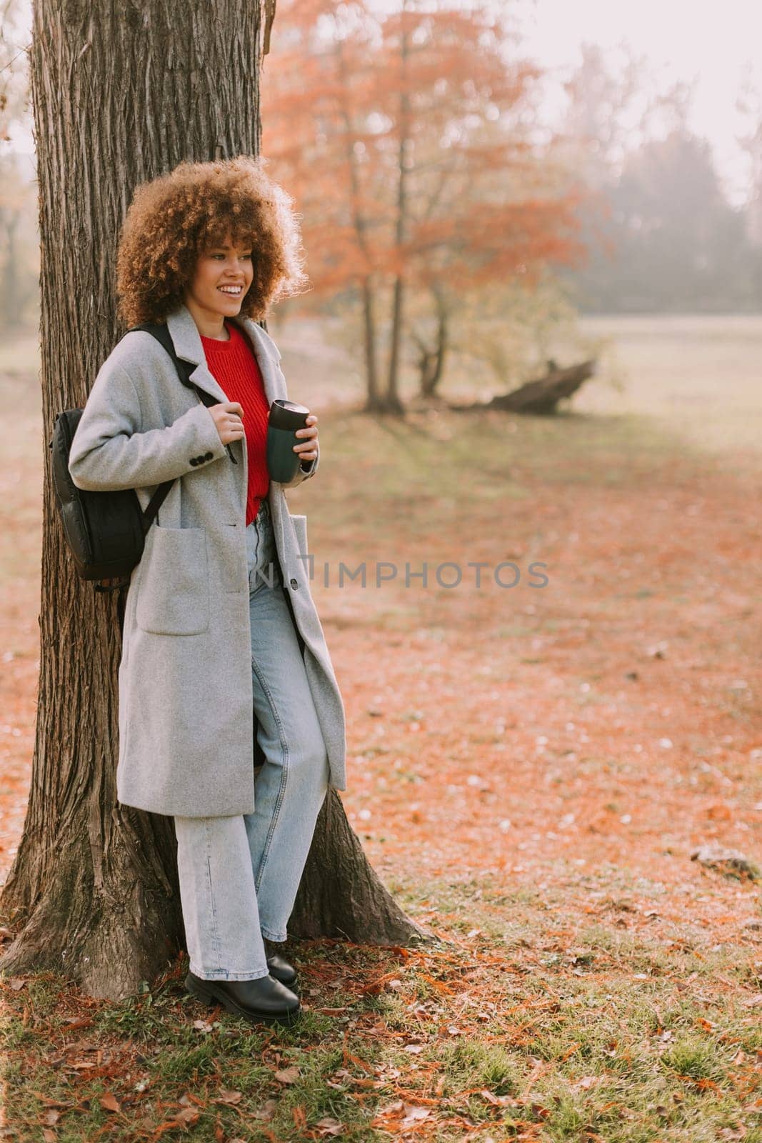 A young woman with curly hair leans against a sturdy tree, holding a warm drink. Dressed in a cozy grey coat, she enjoys the tranquil atmosphere of an autumn day.