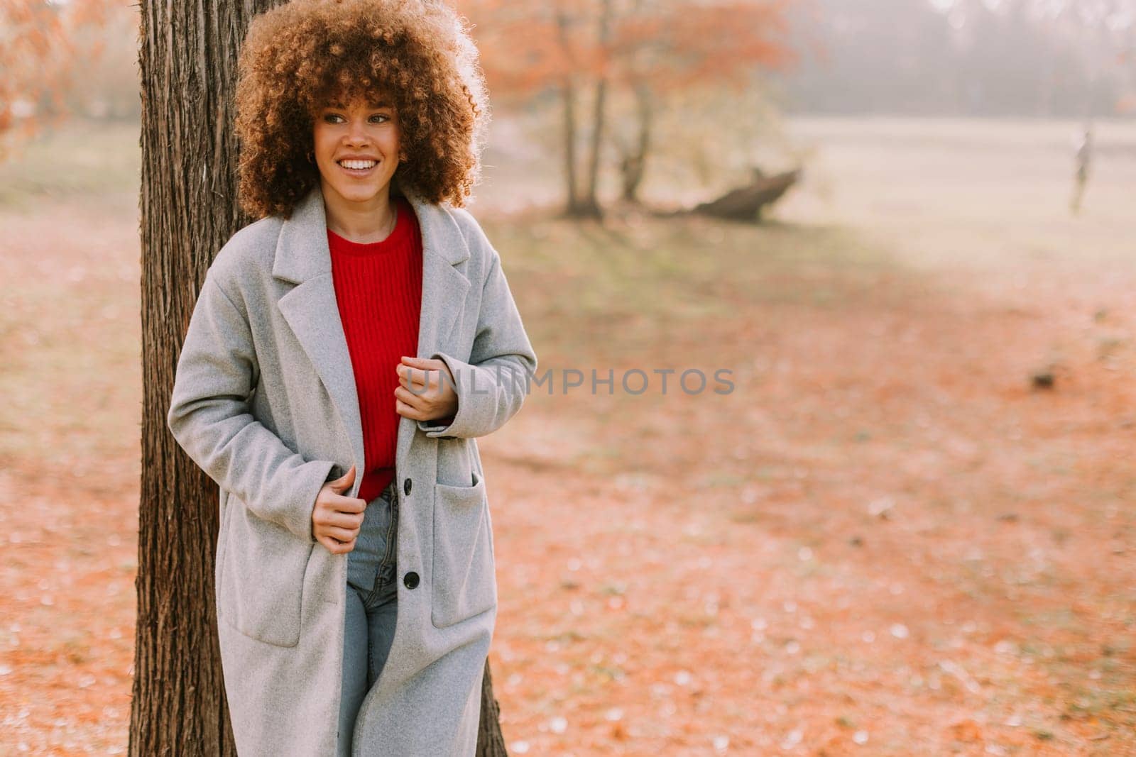 A young woman with curly hair leans against a tree, wearing a warm sweater, smiling joyfully amidst the golden autumn foliage and fallen leaves around her.