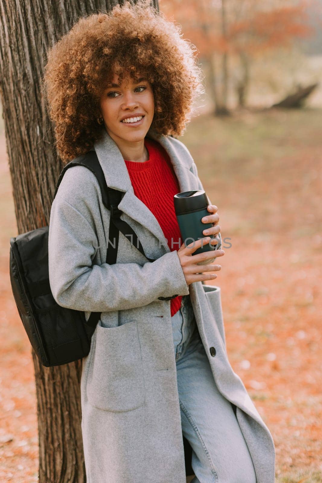A young woman with curly hair leans against a sturdy tree, holding a warm drink. Dressed in a cozy grey coat, she enjoys the tranquil atmosphere of an autumn day.