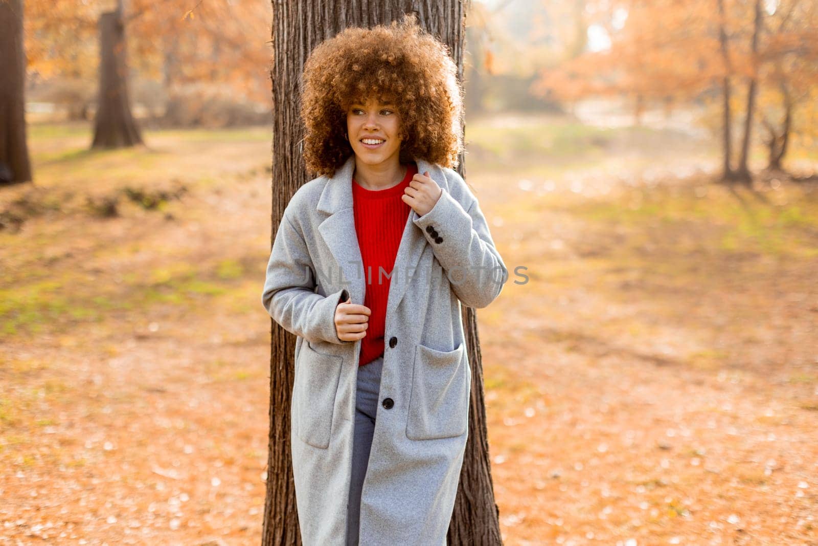 A young woman with curly hair leans against a tree, wearing a warm sweater, smiling joyfully amidst the golden autumn foliage and fallen leaves around her.
