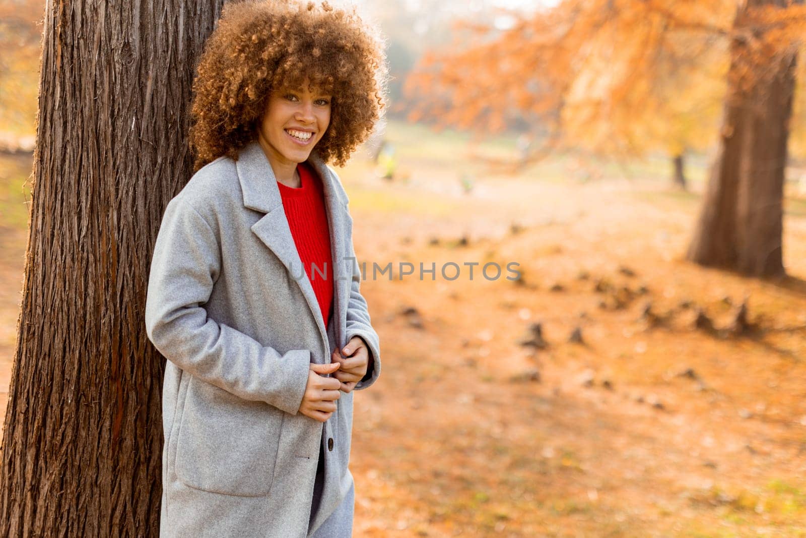 A young woman with curly hair leans against a tree, wearing a warm sweater, smiling joyfully amidst the golden autumn foliage and fallen leaves around her.