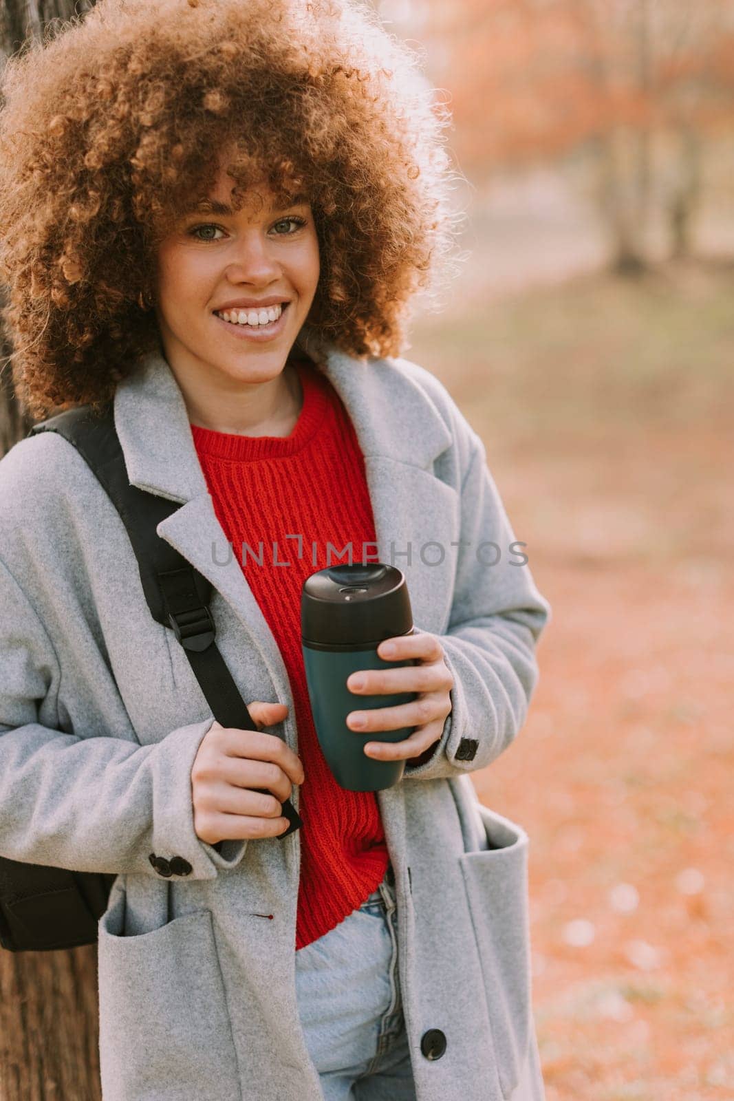 A young woman with curly hair leans against a sturdy tree, holding a warm drink. Dressed in a cozy grey coat, she enjoys the tranquil atmosphere of an autumn day.