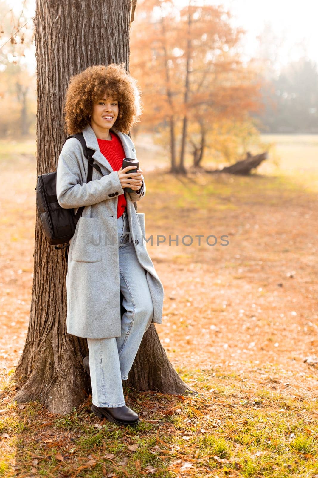 A young woman with curly hair leans against a sturdy tree, holding a warm drink. Dressed in a cozy grey coat, she enjoys the tranquil atmosphere of an autumn day.