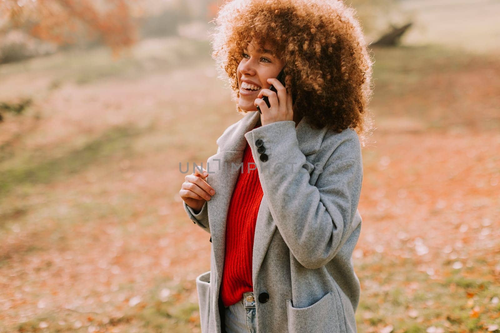 A young woman with curly hair laughs while talking on her phone, dressed in a stylish gray coat and vibrant red sweater, surrounded by the warm hues of autumn foliage.