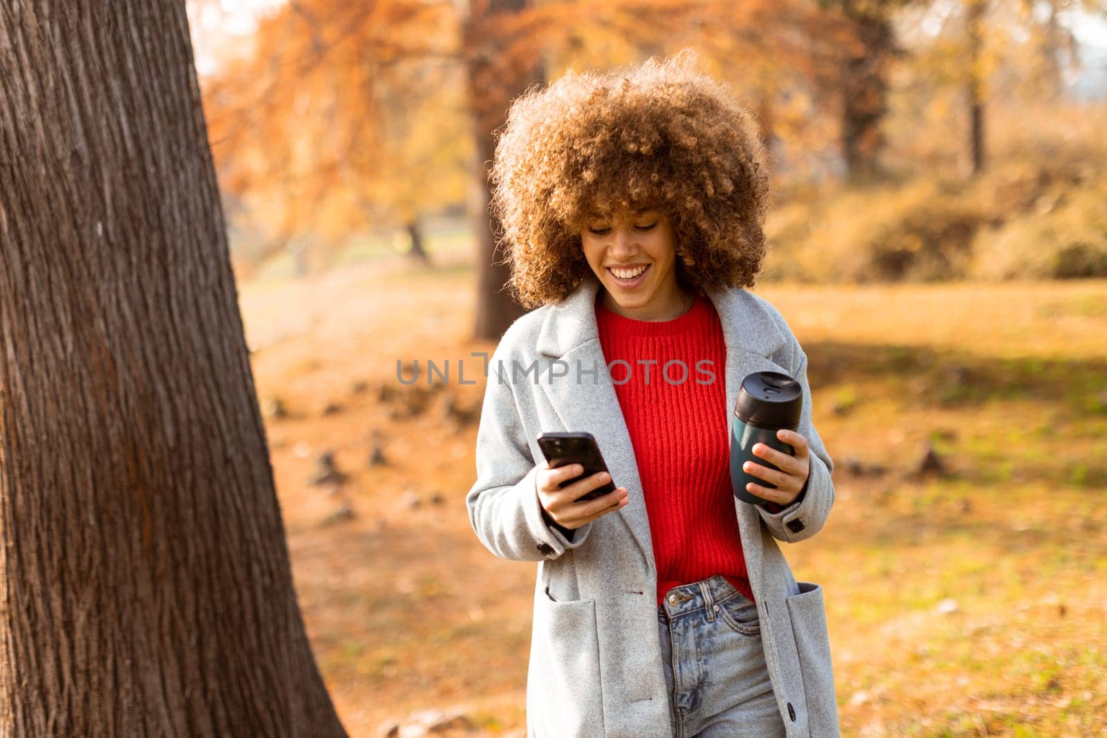 A young woman with curly hair wears a cozy sweater and a stylish coat while walking through a vibrant autumn park, happily engaged with her phone and coffee.