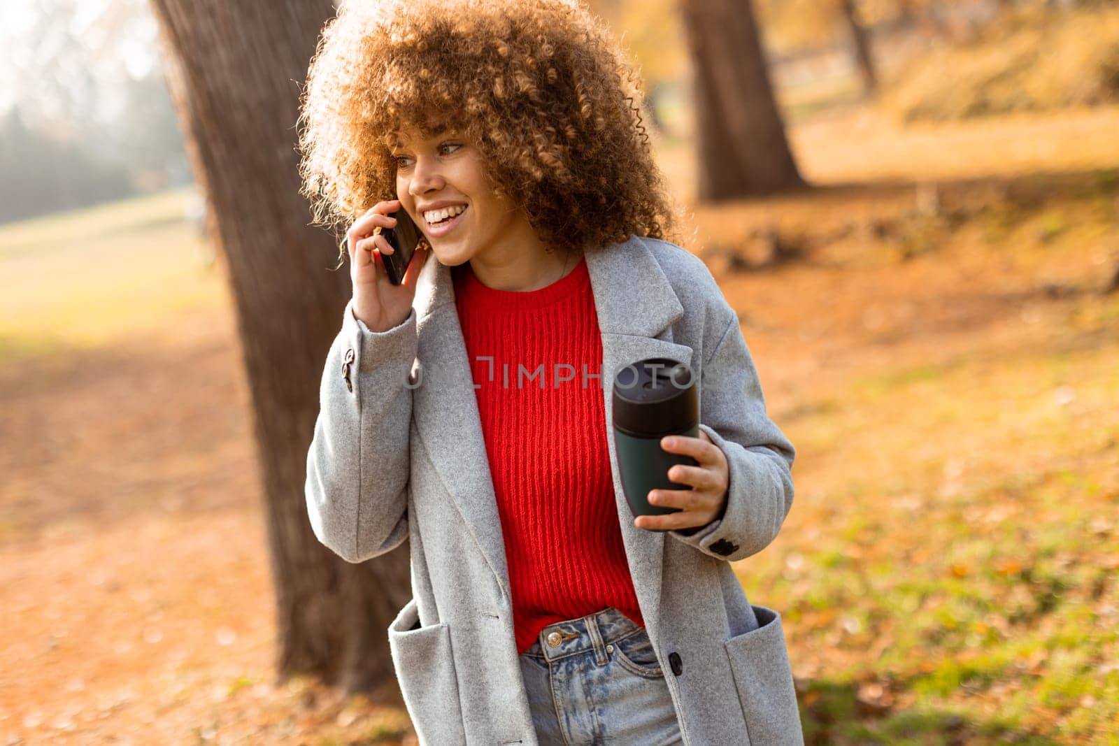 Bright autumn foliage surrounds a young woman, who is smiling and conversing on her phone, with a warm beverage in hand. She embodies a joyful moment in nature.