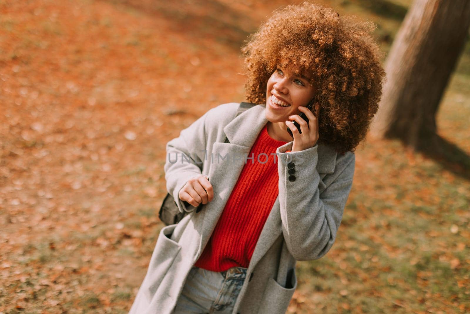A young woman with curly hair laughs while talking on her phone, dressed in a stylish gray coat and vibrant red sweater, surrounded by the warm hues of autumn foliage.