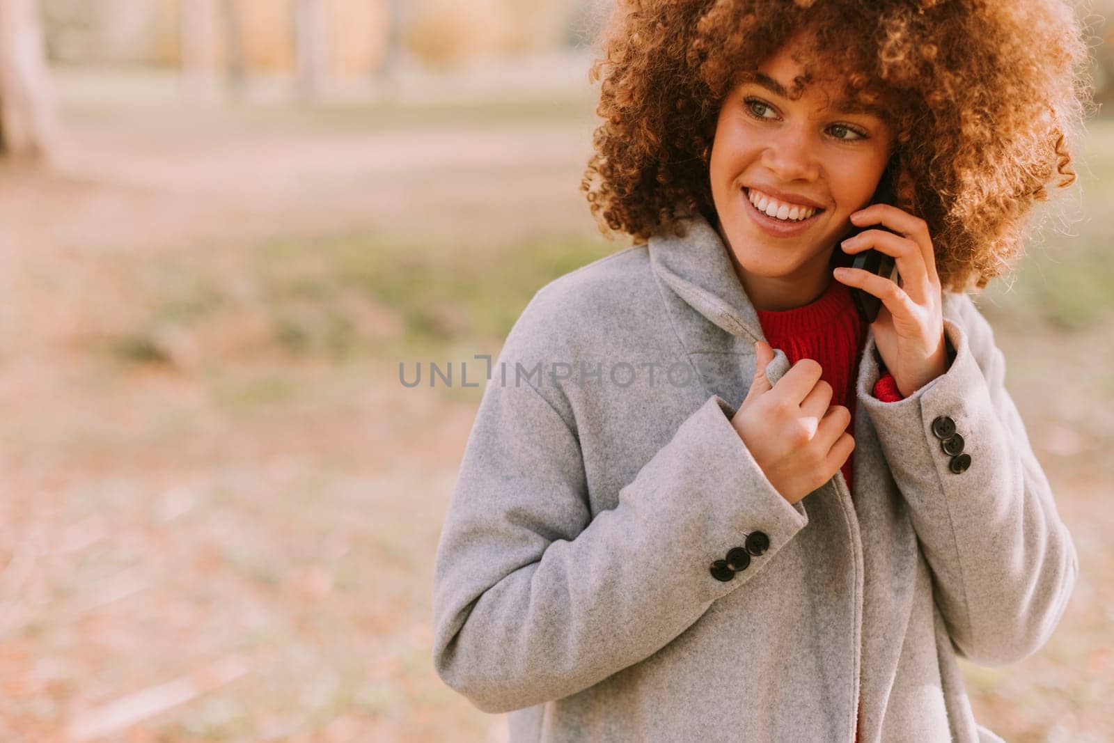 A young woman with curly hair smiles brightly while talking on her phone, dressed warmly in a stylish coat amidst a picturesque autumn landscape filled with soft light.