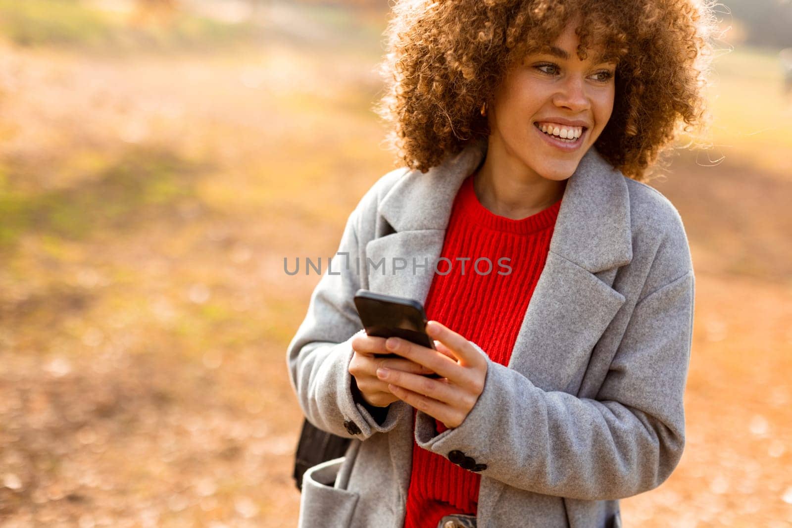 A cheerful individual with curly hair smiles brightly as she engages with her smartphone, wearing a cozy red sweater and a stylish gray coat amidst golden autumn foliage.
