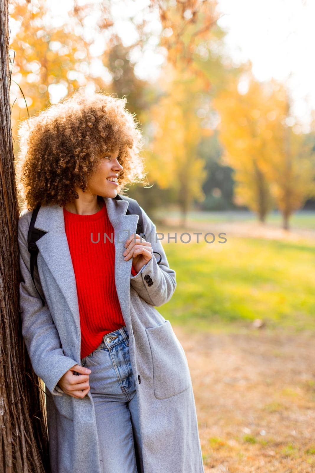 A joyful woman with curly hair leans against a tree in a park filled with golden leaves. Dressed in a red sweater and gray coat, she embodies the essence of autumn bliss.