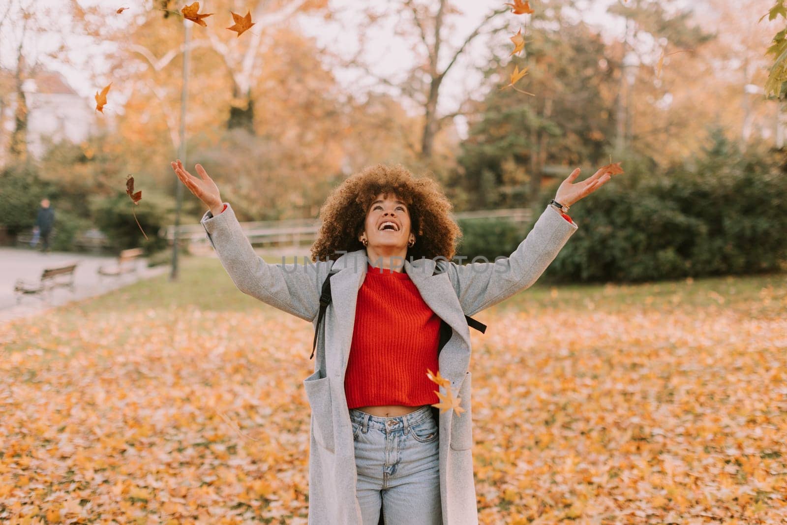 A cheerful individual with curly hair plays in a park during autumn, joyfully tossing vibrant leaves into the air among a golden blanket of fallen foliage.