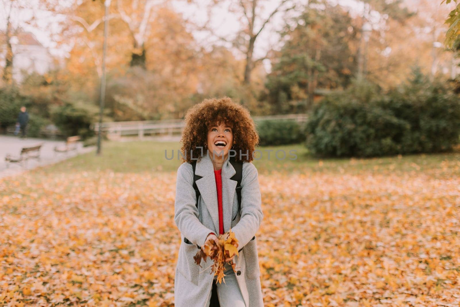 A cheerful individual with curly hair plays in a park during autumn, joyfully tossing vibrant leaves into the air among a golden blanket of fallen foliage.