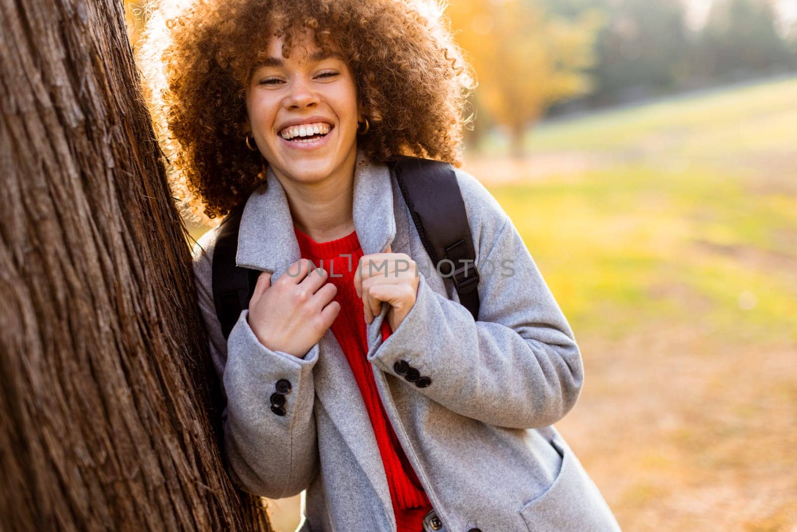 A carefree young woman laughs heartily while standing next to a tree in a serene park. The warm sunlight filters through the leaves, creating a joyful atmosphere.