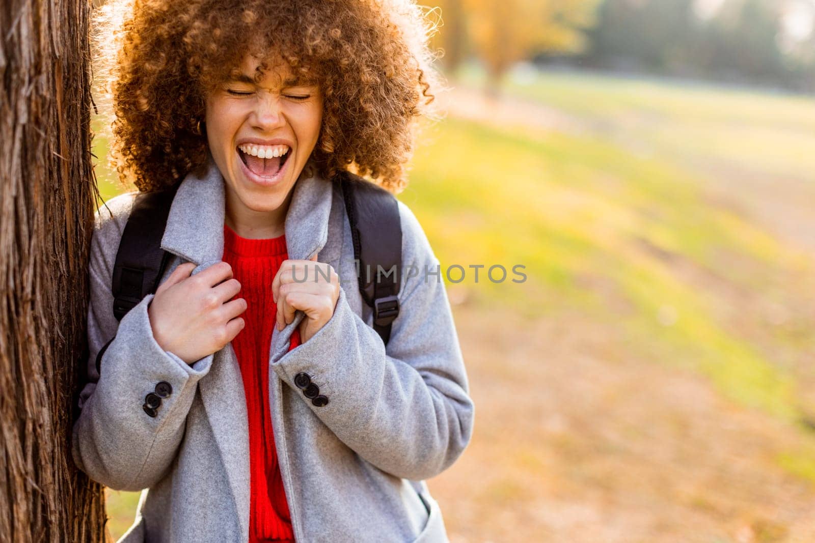 A carefree young woman laughs heartily while standing next to a tree in a serene park. The warm sunlight filters through the leaves, creating a joyful atmosphere.