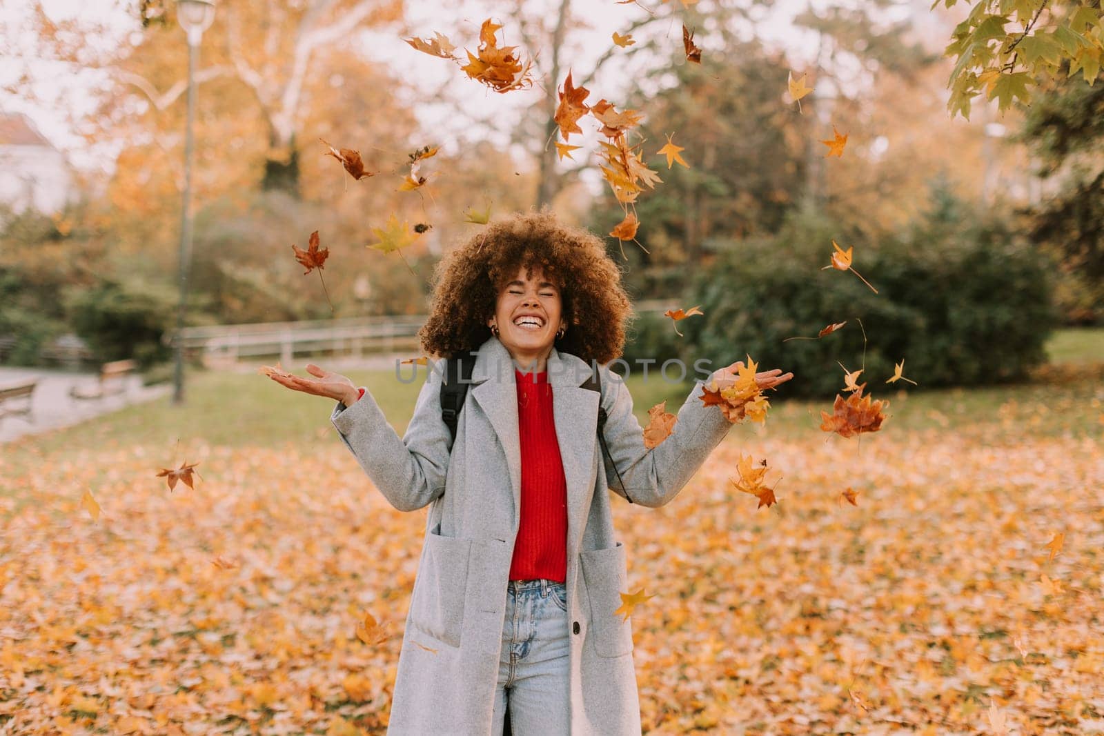 A cheerful individual with curly hair plays in a park during autumn, joyfully tossing vibrant leaves into the air among a golden blanket of fallen foliage.