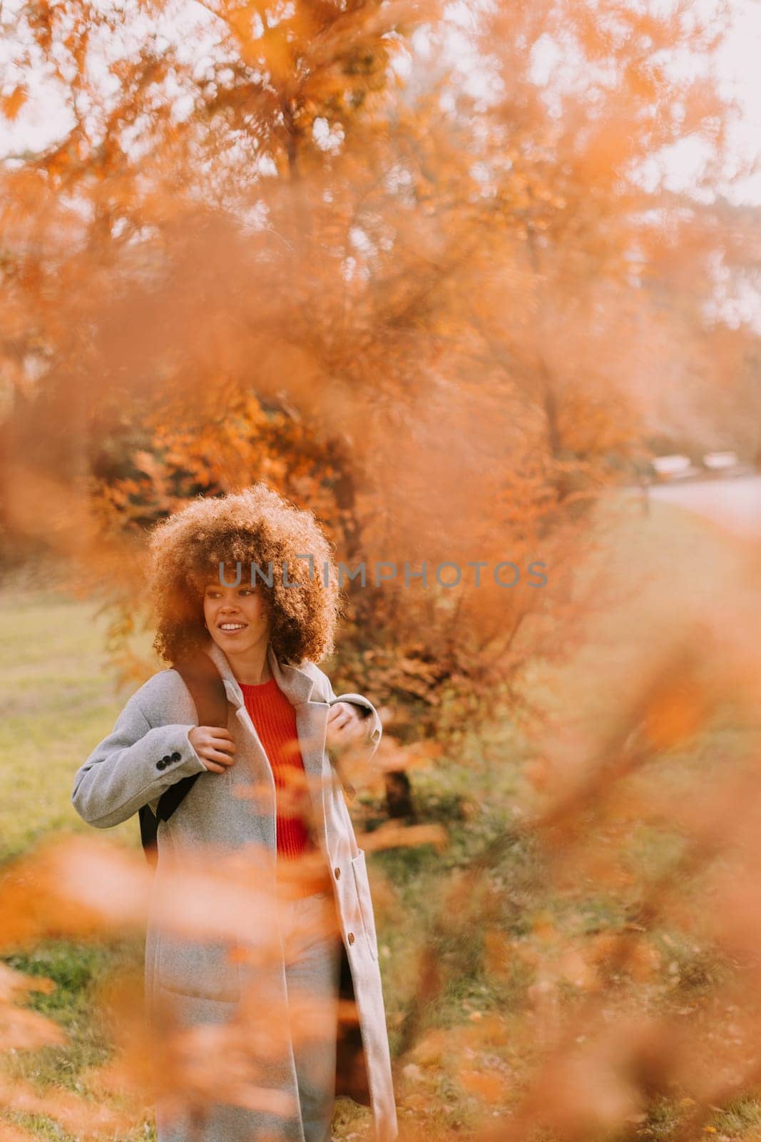 In a serene park filled with stunning autumn colors, a joyful woman stands with a bright smile, embracing the beauty of nature as leaves fall gently around her.