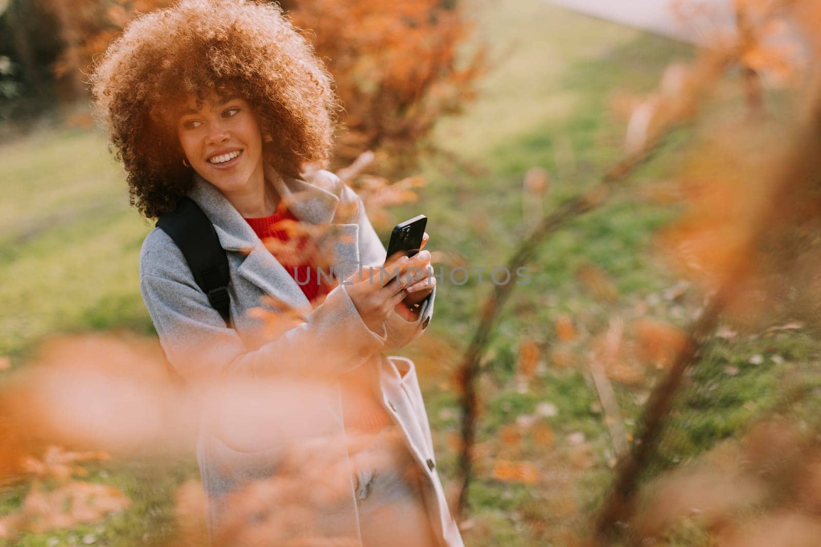 A joyful person with curly hair smiles while holding a camera, surrounded by vibrant autumn leaves in a serene outdoor setting on a sunny day.