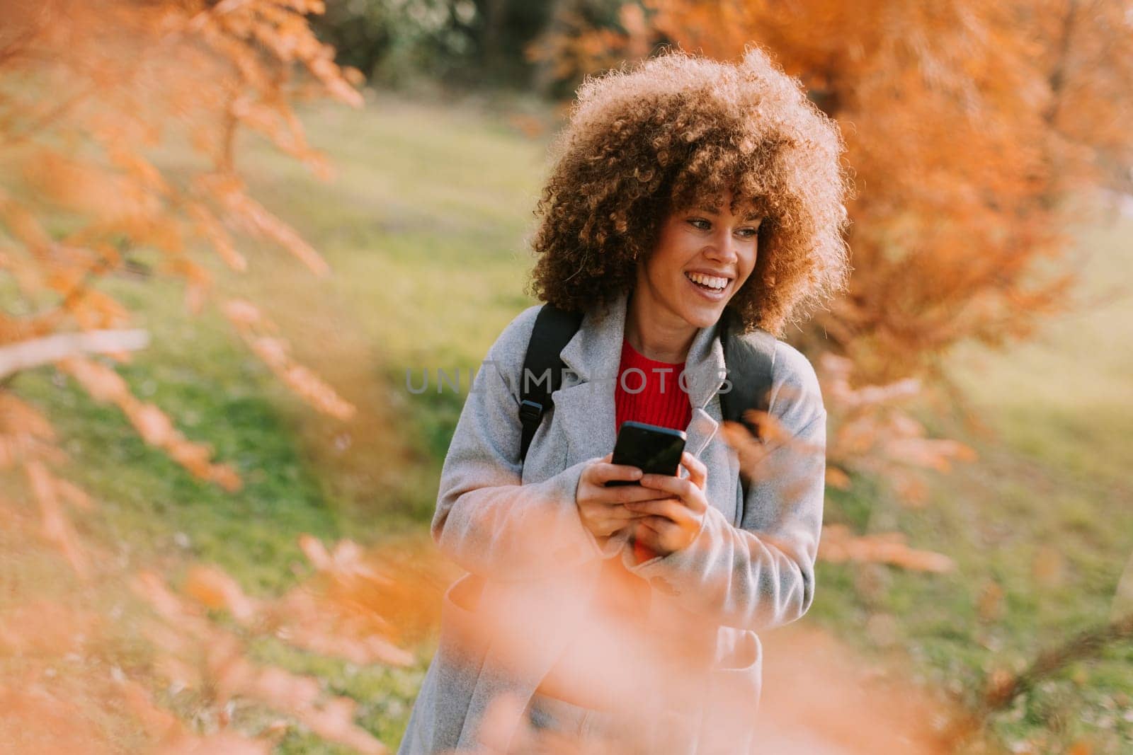 A joyful person with curly hair smiles while holding a camera, surrounded by vibrant autumn leaves in a serene outdoor setting on a sunny day.