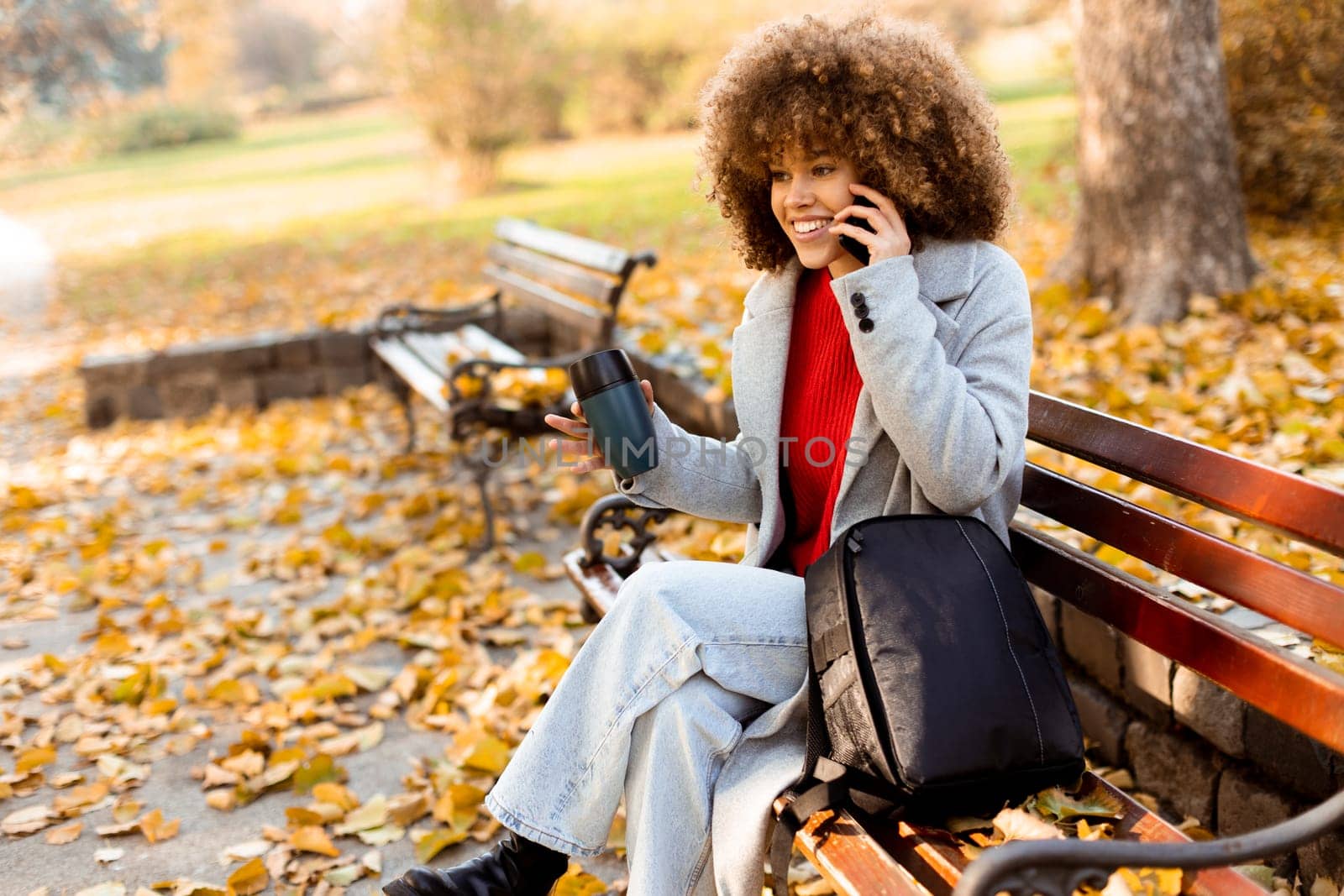 Sitting comfortably on a bench, a cheerful young woman with curly hair talks on her phone while enjoying a cup of coffee amidst fallen autumn leaves in the park.