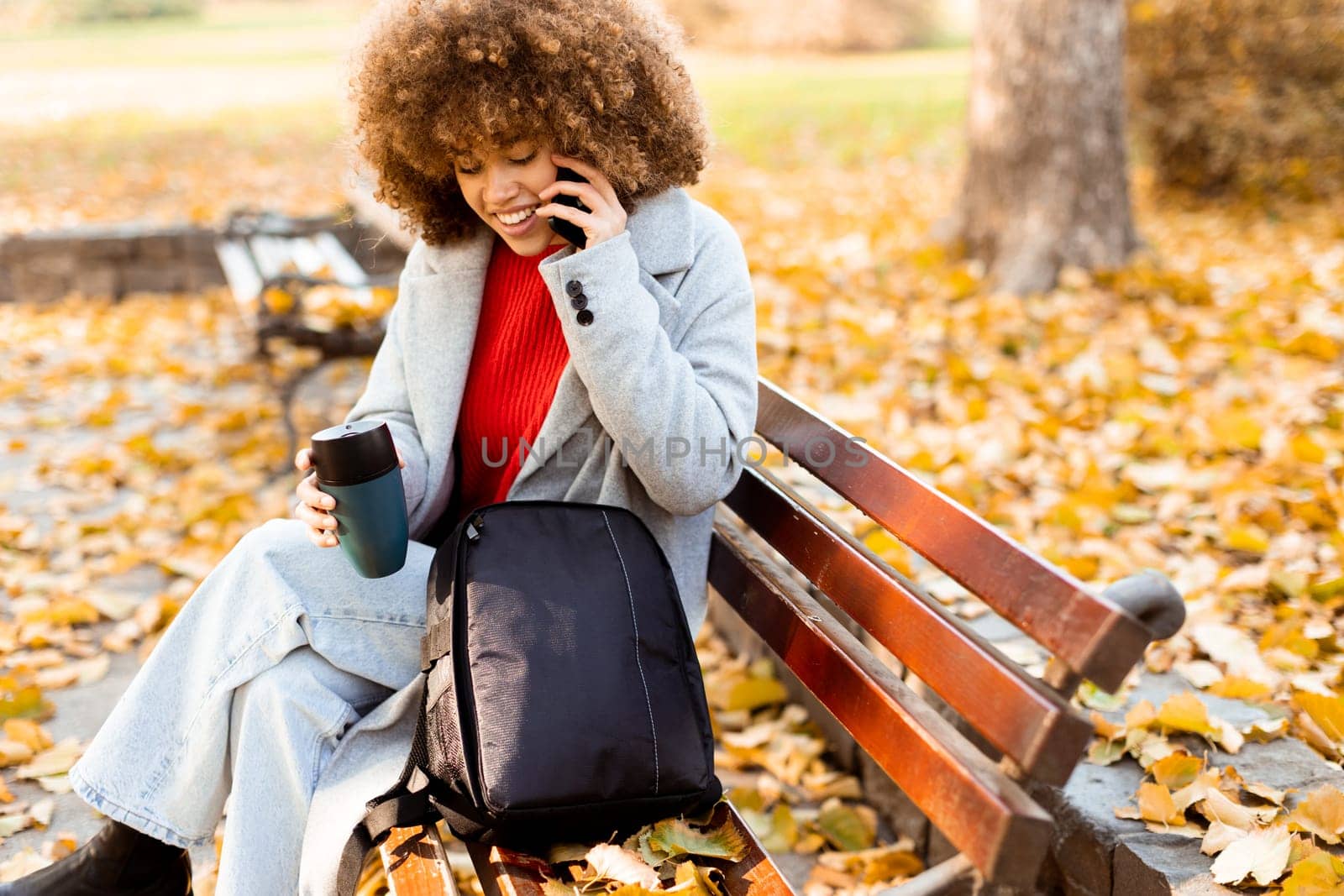 Sitting comfortably on a bench, a cheerful young woman with curly hair talks on her phone while enjoying a cup of coffee amidst fallen autumn leaves in the park.