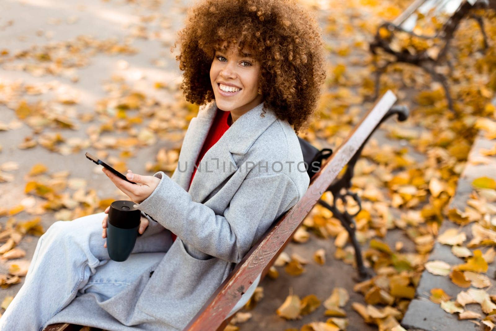 Sitting on a wooden bench in a park, a young woman smiles while texting on her smartphone, enjoying the vibrant autumn leaves that blanket the ground.