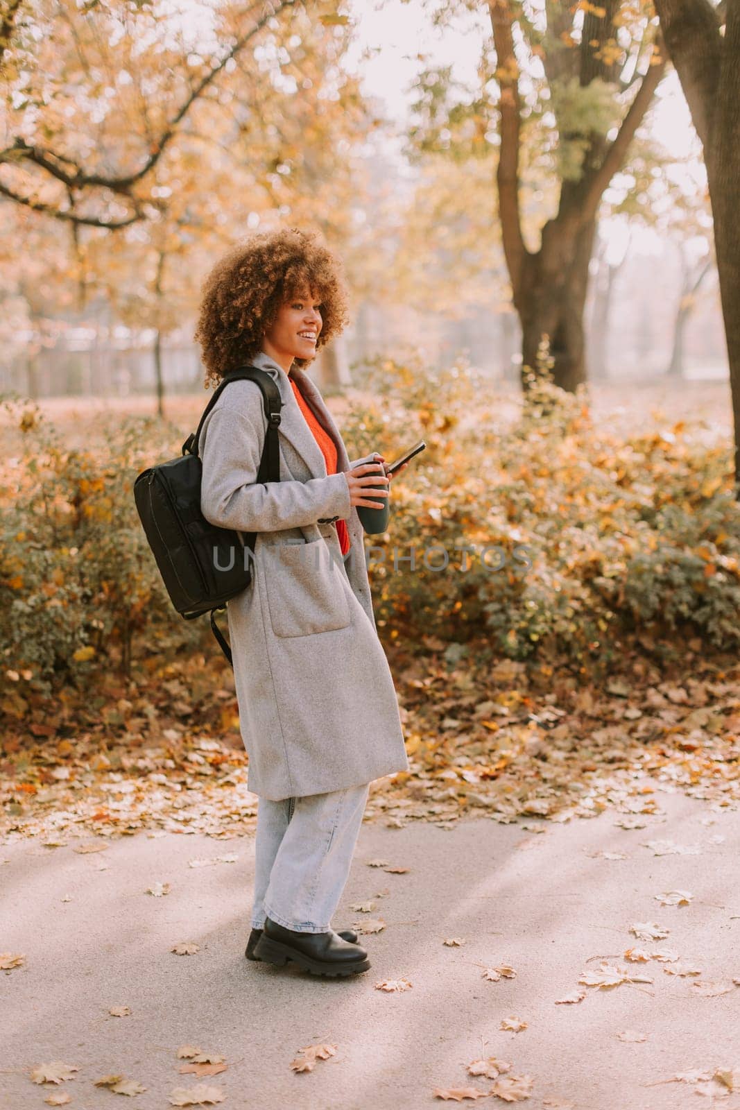 Joyful woman stands amidst vibrant autumn foliage in a park, sipping a warm drink and wearing a cozy outfit, embodying the beauty of the season in the afternoon light.