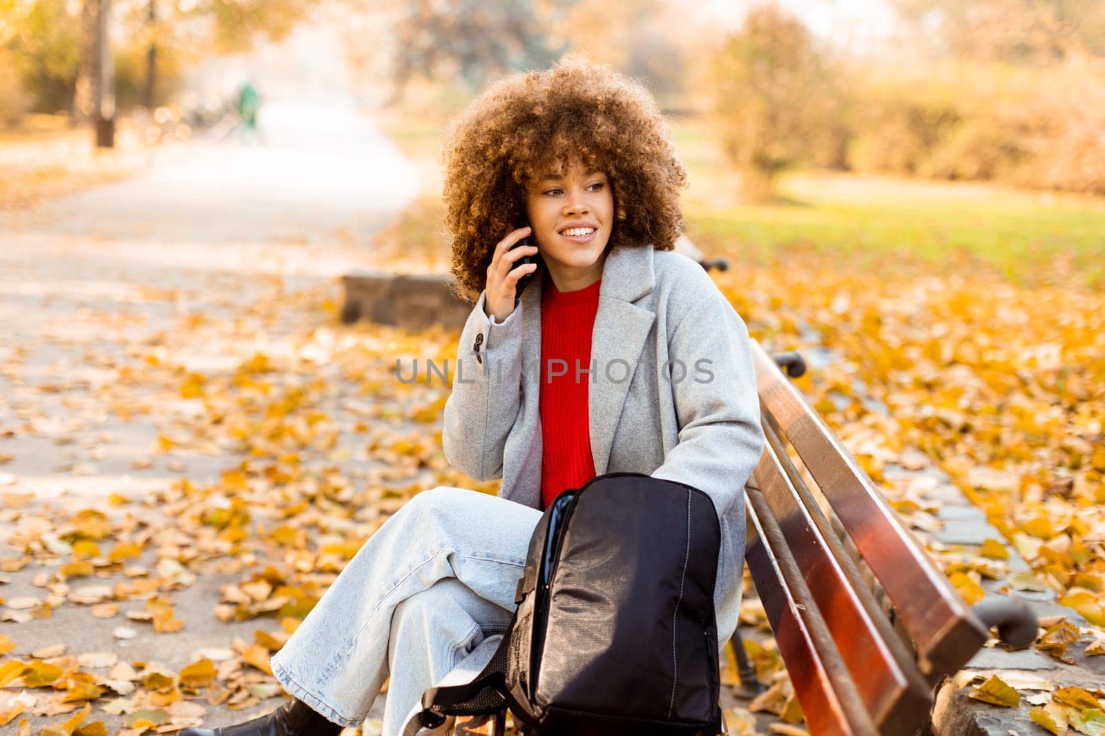 A joyful woman with curly hair makes a phone call while sitting on a park bench surrounded by vibrant autumn foliage, enjoying a beautiful day outdoors.