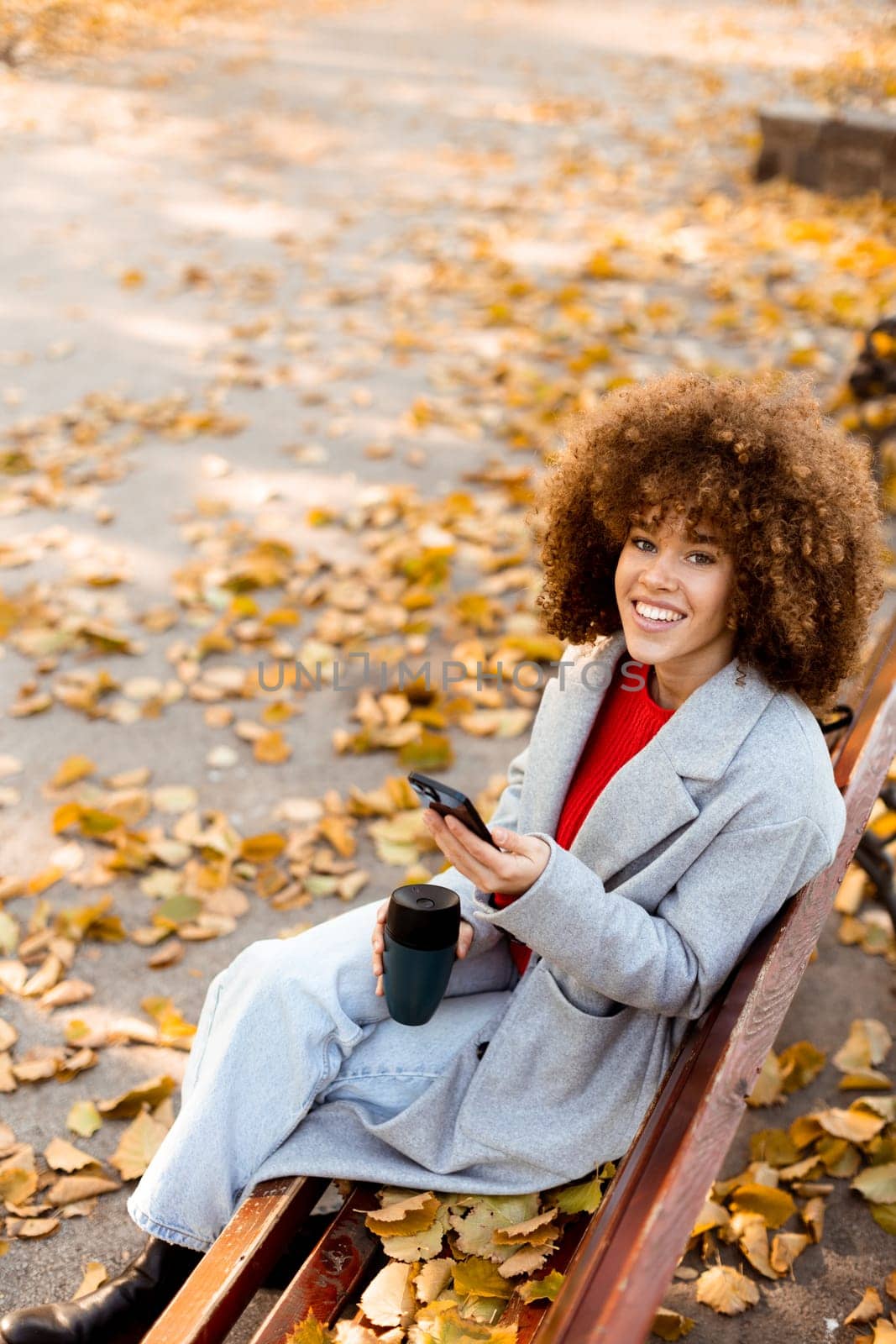 Sitting on a wooden bench in a park, a young woman smiles while texting on her smartphone, enjoying the vibrant autumn leaves that blanket the ground.