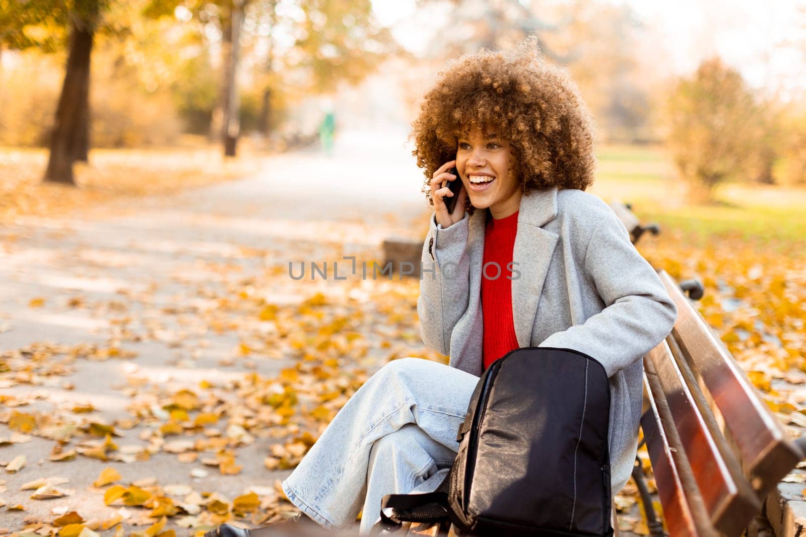 A joyful woman with curly hair makes a phone call while sitting on a park bench surrounded by vibrant autumn foliage, enjoying a beautiful day outdoors.