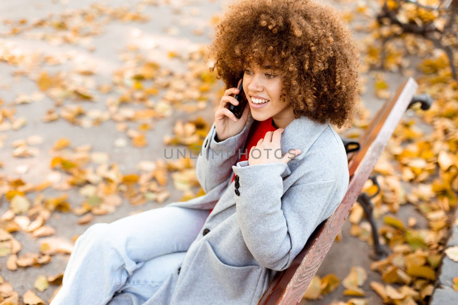 A young woman with curly hair laughs joyfully while talking on her phone, sitting on a park bench covered in vibrant autumn leaves