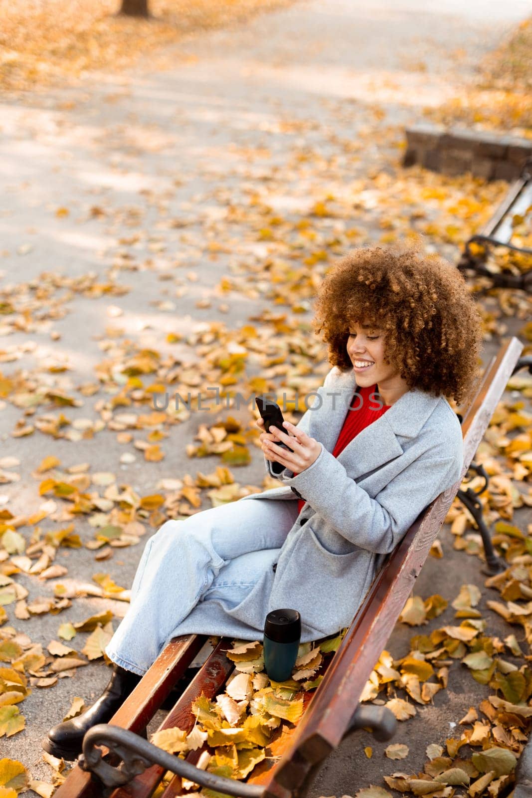 Sitting on a wooden bench in a park, a young woman smiles while texting on her smartphone, enjoying the vibrant autumn leaves that blanket the ground.