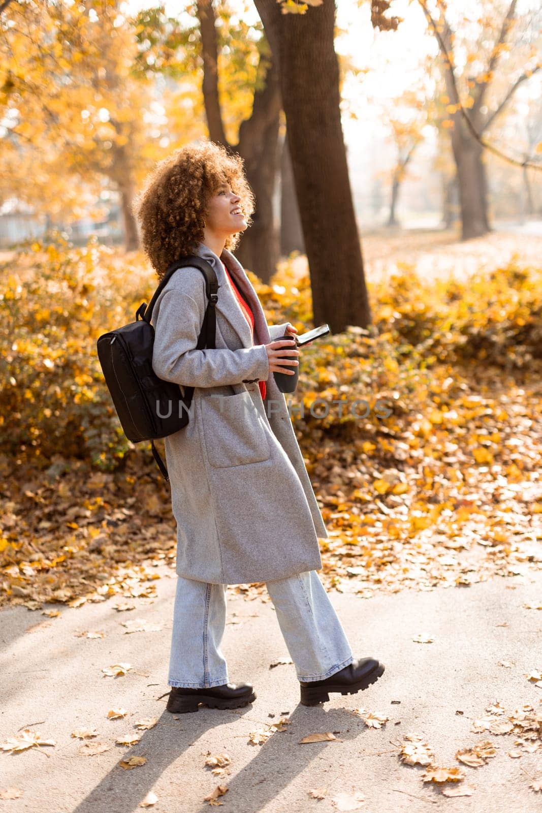 Joyful woman stands amidst vibrant autumn foliage in a park, sipping a warm drink and wearing a cozy outfit, embodying the beauty of the season in the afternoon light.
