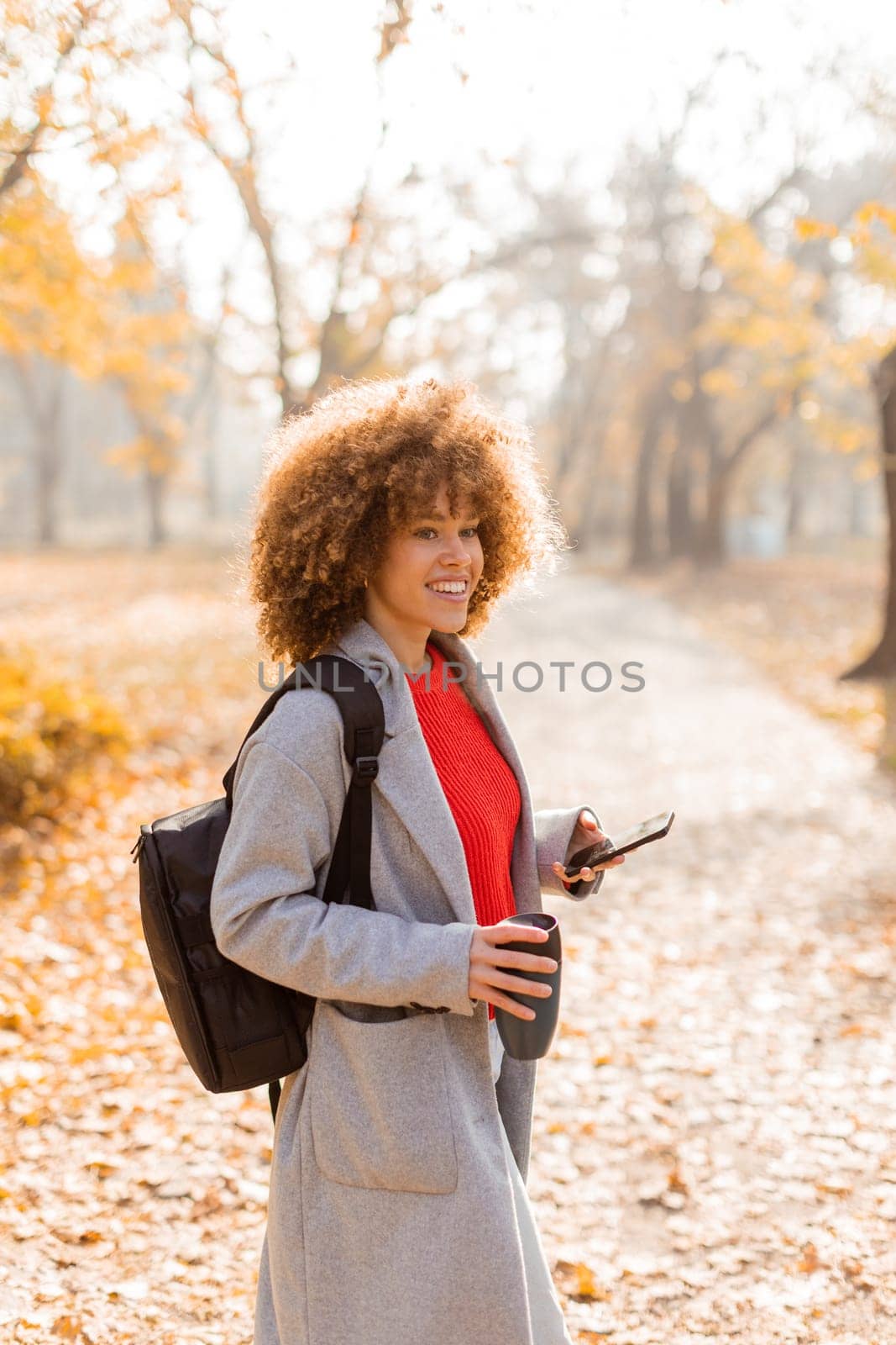 Joyful woman stands amidst vibrant autumn foliage in a park, sipping a warm drink and wearing a cozy outfit, embodying the beauty of the season in the afternoon light.