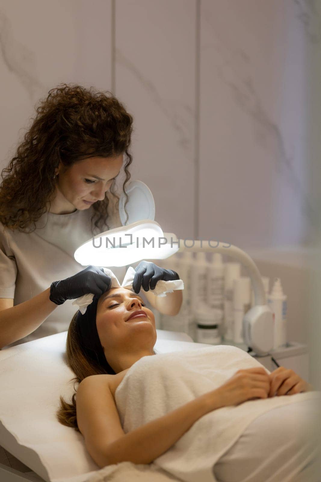 A woman enjoys a rejuvenating facial treatment at a tranquil spa. A skilled aesthetician gently wipes her forehead, enhancing her pampering experience.
