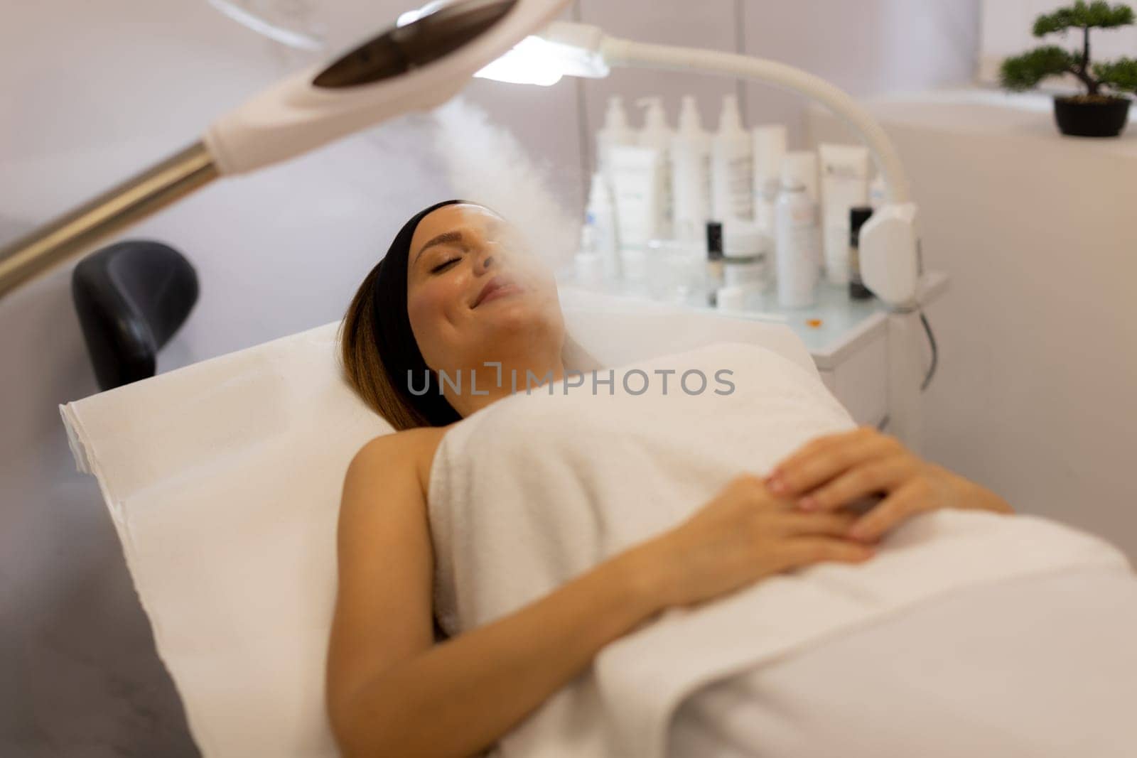 A woman comfortably reclines on a treatment bed, enjoying a calming facial steam at a serene spa. Soft light enhances the peaceful, rejuvenating atmosphere.