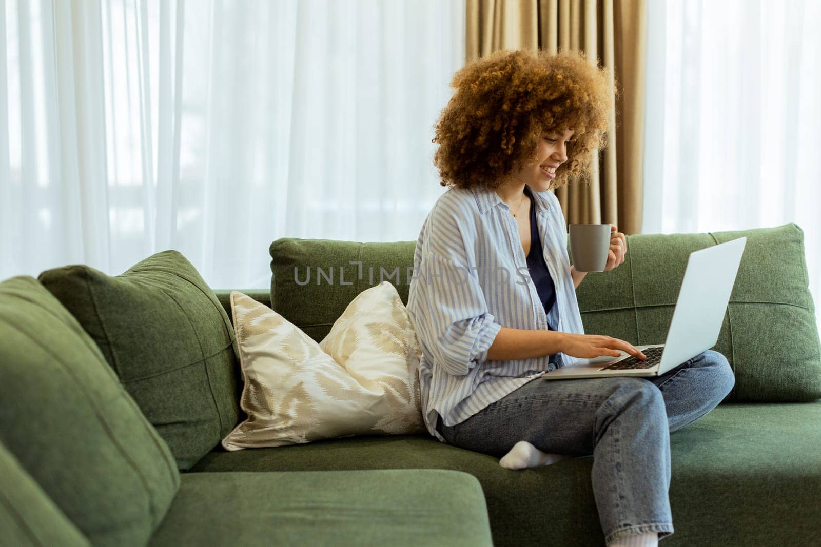 Sitting comfortably on a plush sofa, a young woman savors her warm coffee while working on her laptop, illuminated by soft natural light in a stylish living space