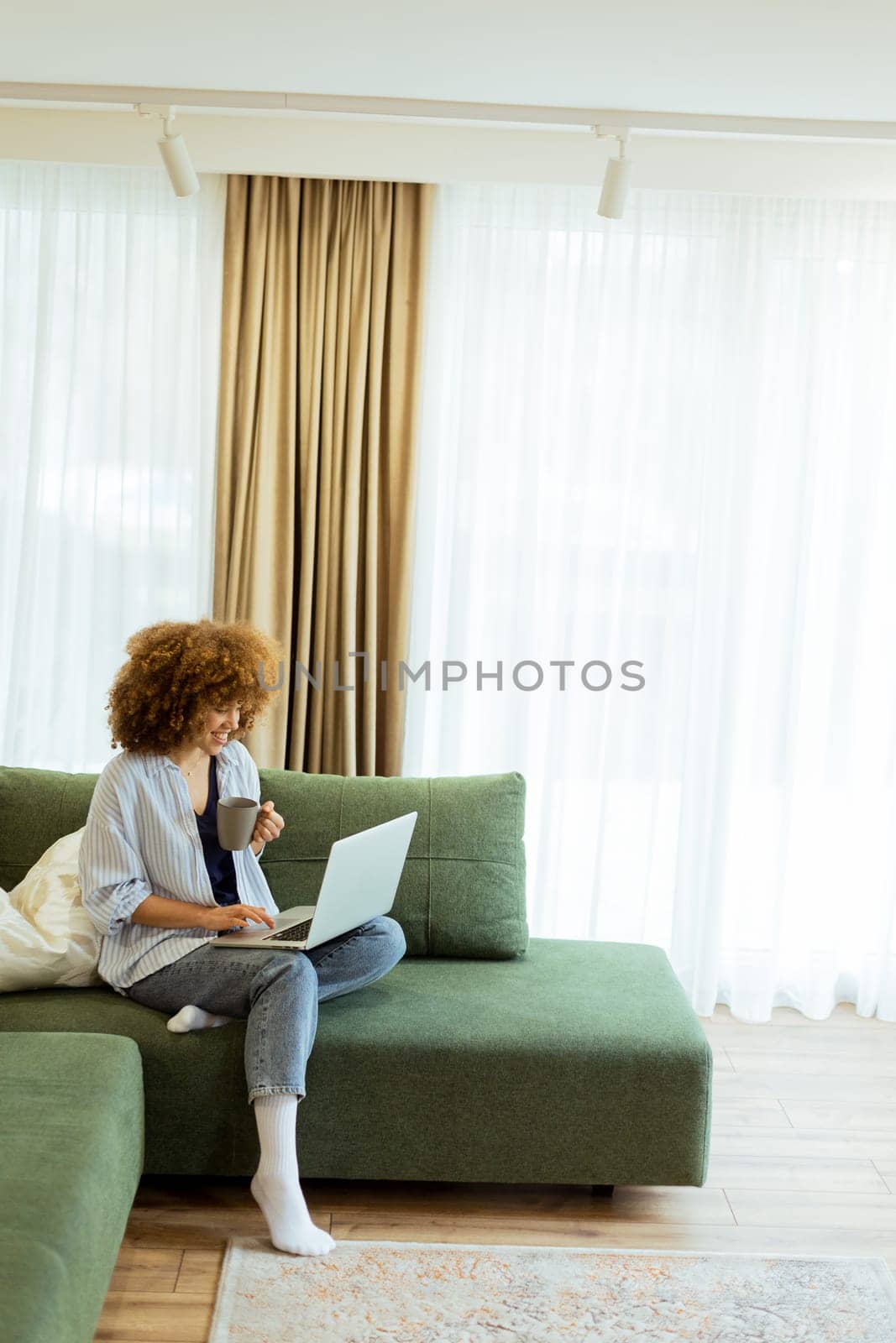 Sitting comfortably on a plush sofa, a young woman savors her warm coffee while working on her laptop, illuminated by soft natural light in a stylish living space