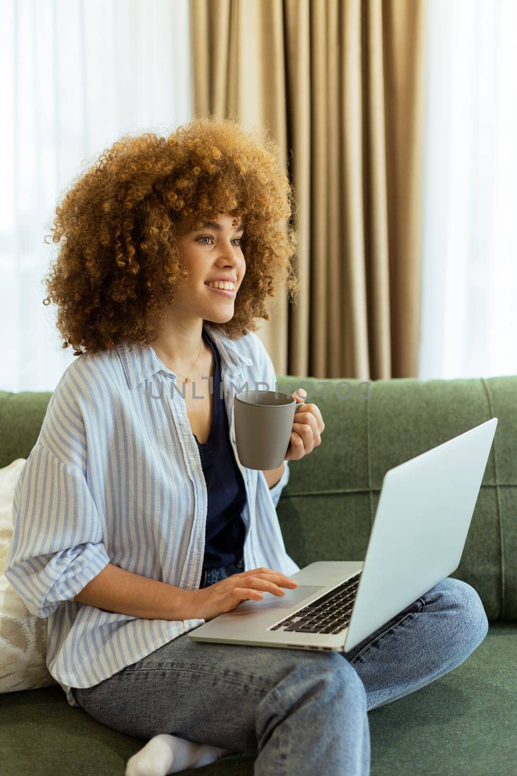 Sitting comfortably on a plush sofa, a young woman savors her warm coffee while working on her laptop, illuminated by soft natural light in a stylish living space