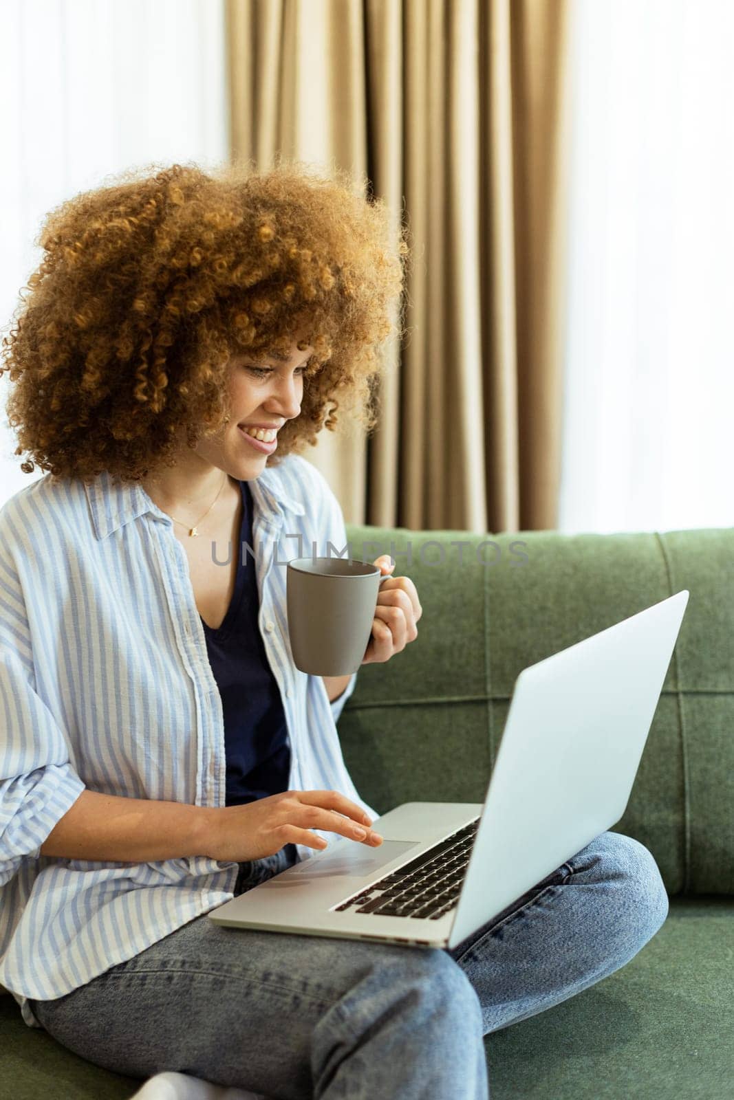 Sitting comfortably on a plush sofa, a young woman savors her warm coffee while working on her laptop, illuminated by soft natural light in a stylish living space