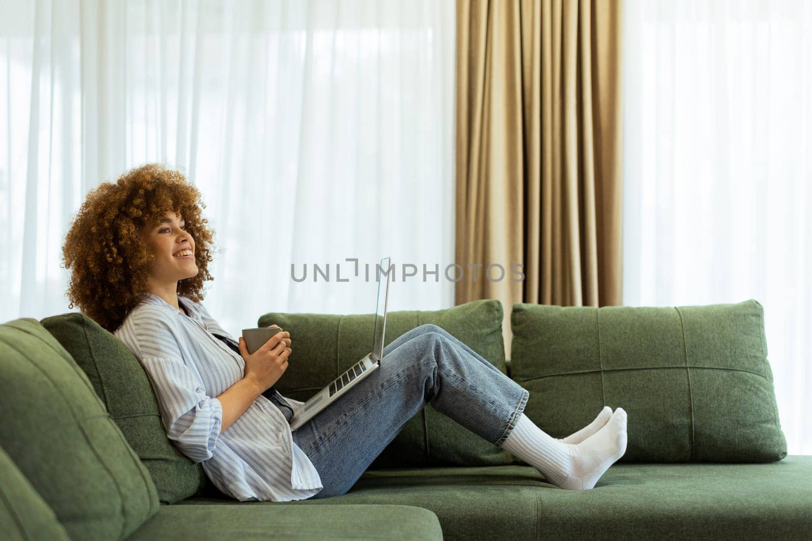 Sitting comfortably on a plush sofa, a young woman savors her warm coffee while working on her laptop, illuminated by soft natural light in a stylish living space