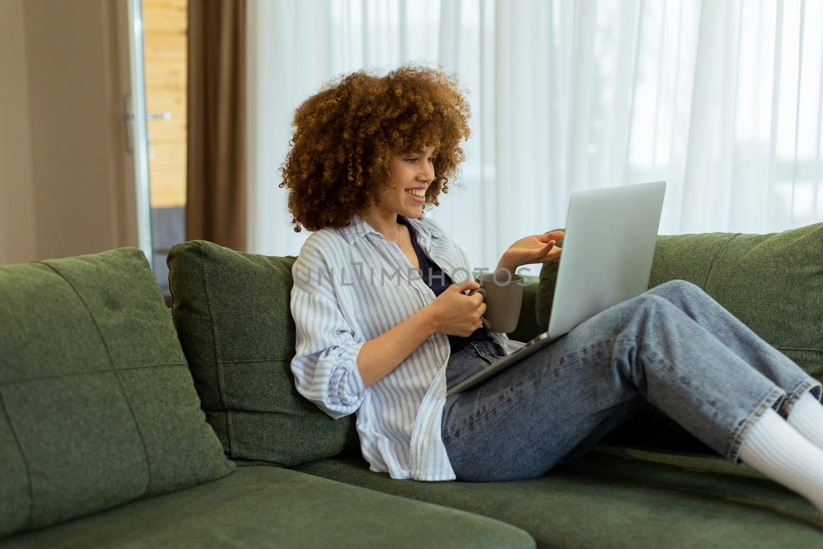 Sitting comfortably on a plush sofa, a young woman savors her warm coffee while working on her laptop, illuminated by soft natural light in a stylish living space