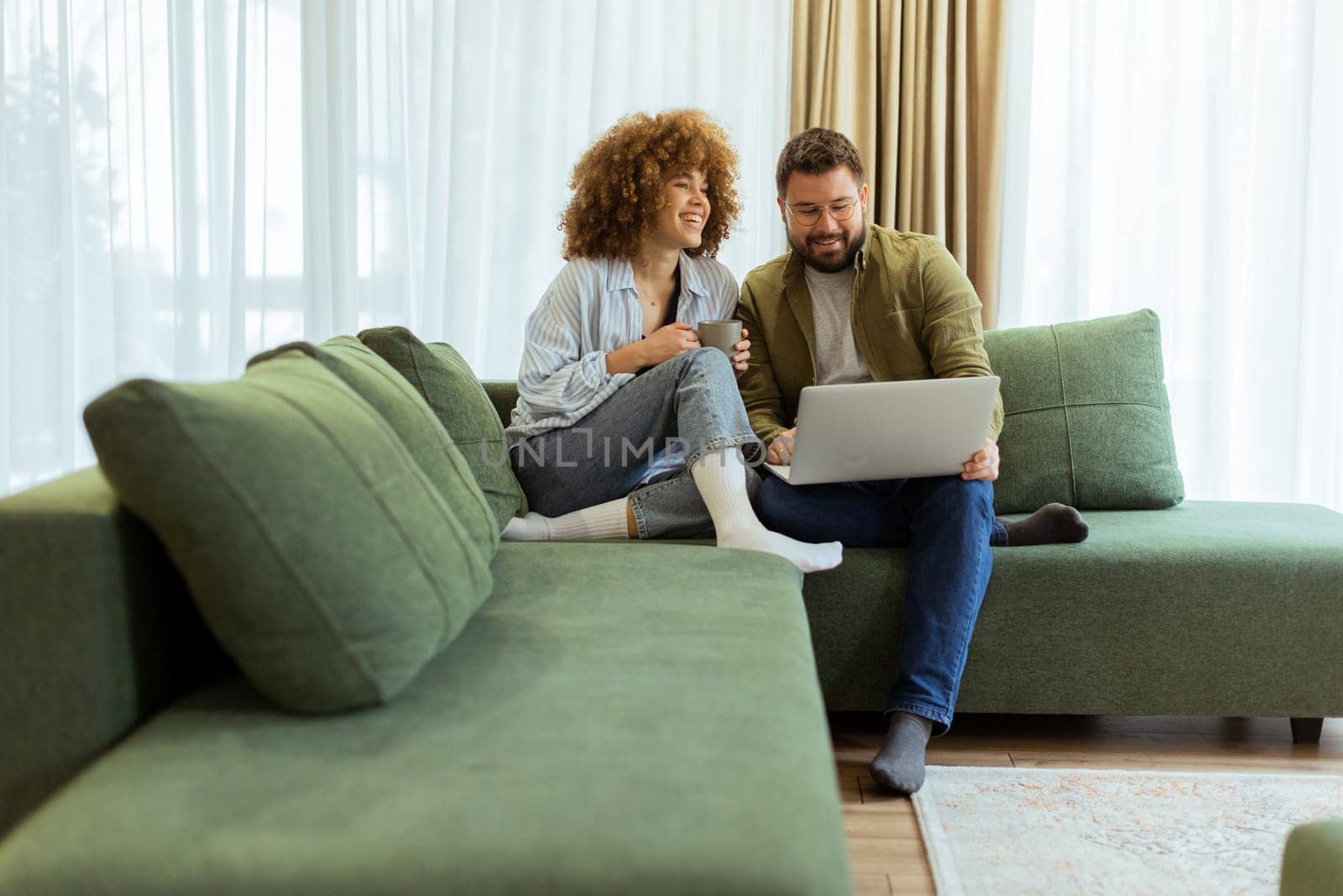 A cheerful couple sits side by side on a plush green couch, engaged in an exciting conversation as they browse through ideas on a laptop under warm sunlight streaming in