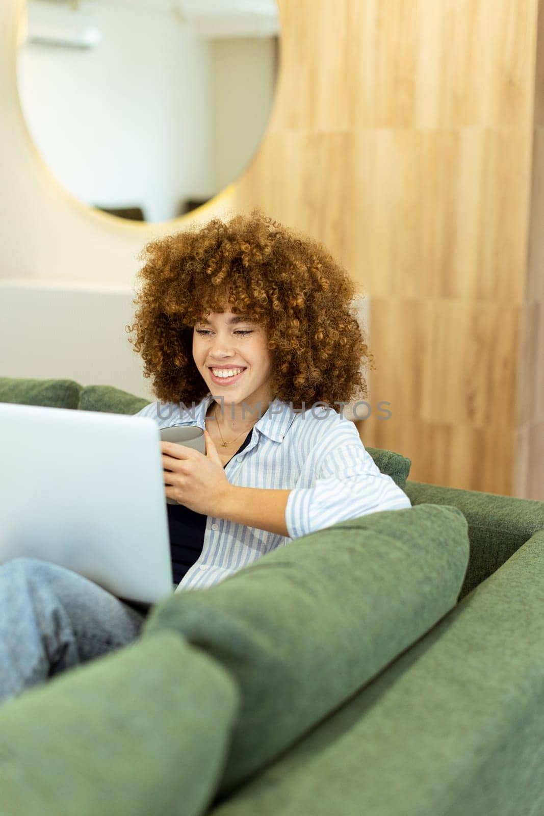 In a bright, modern living area, a young woman with curly hair smiles while using her laptop, surrounded by soft green cushions. Sunlight filters in, creating a warm atmosphere.