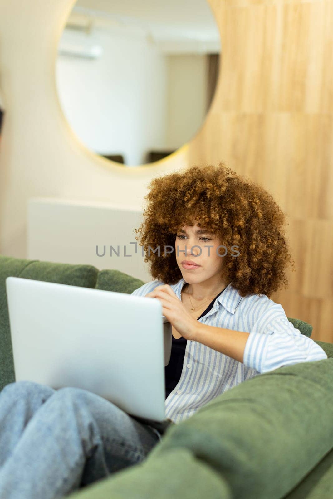 A young woman with curly hair is absorbed in her work on a laptop, seated comfortably on a plush green sofa in a contemporary indoor environment illuminated by soft lighting.