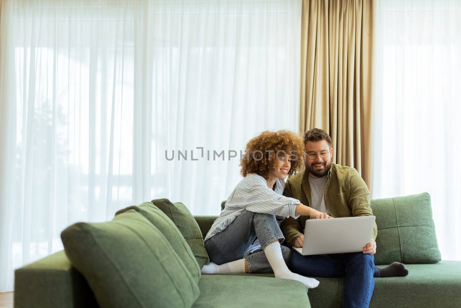 Friends share a joyful moment on a green sofa, discussing ideas while one enjoys a warm drink and the other works on a laptop, surrounded by soft natural light.