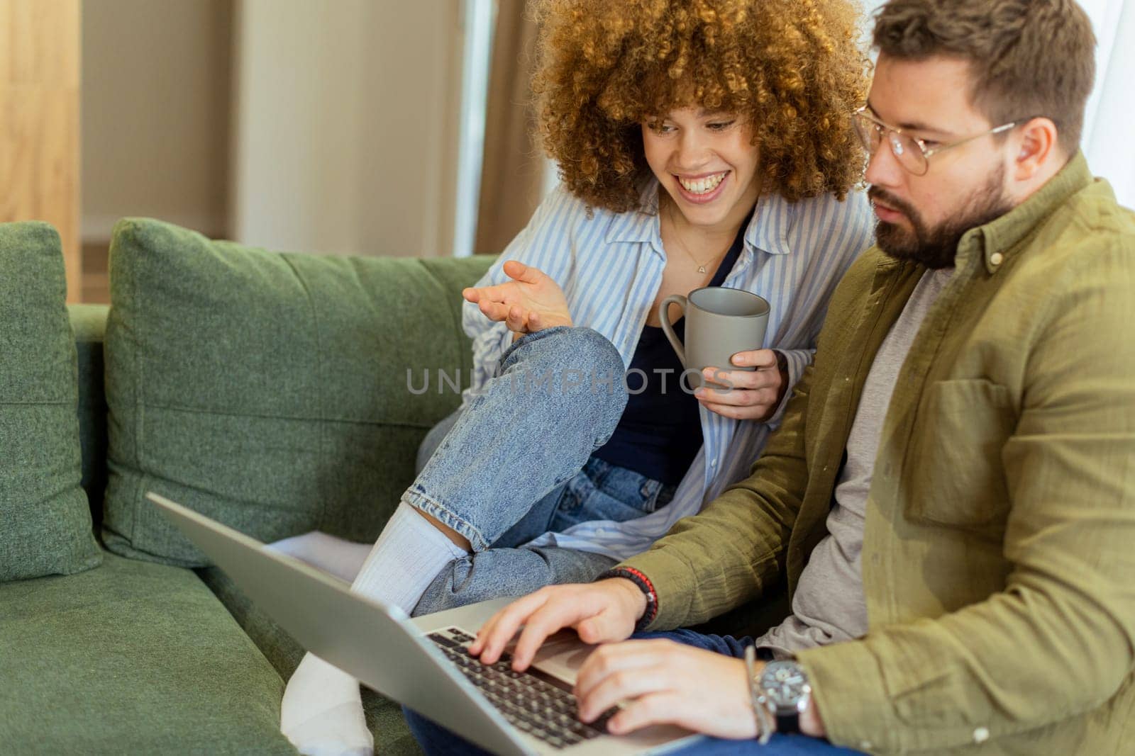 In a stylish living room, a young couple shares a joyful moment as they collaborate on a laptop, surrounded by soft green furnishings and warm sunlight streaming through the windows.