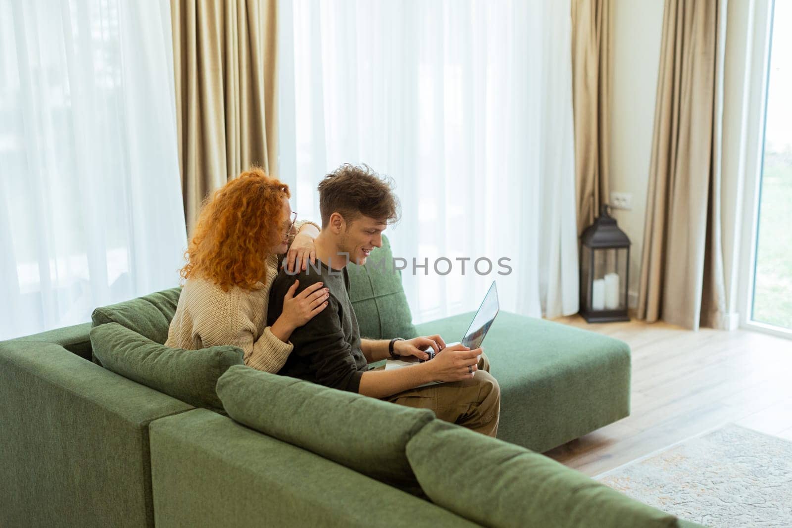 Two friends engage in a light-hearted moment at home, seated comfortably on a green couch, as they collaborate on a laptop, filled with laughter and joy.