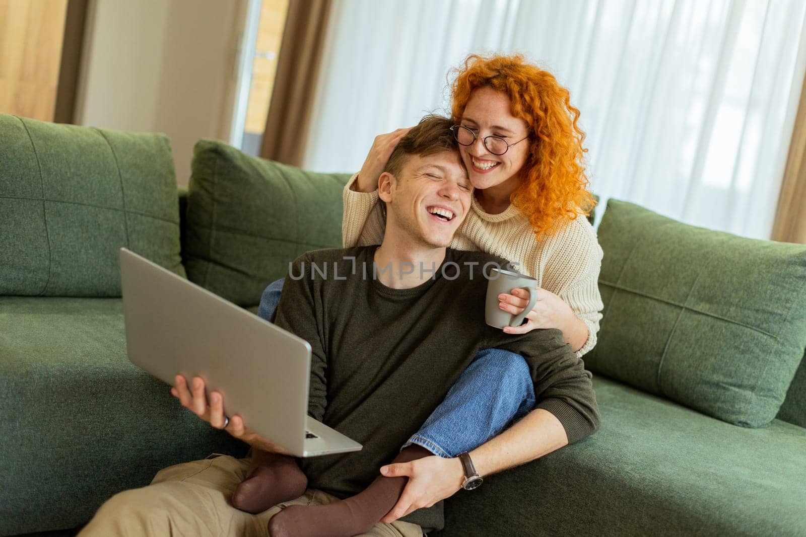Two friends engage in a light-hearted moment at home, seated comfortably on a green couch, as they collaborate on a laptop, filled with laughter and joy.