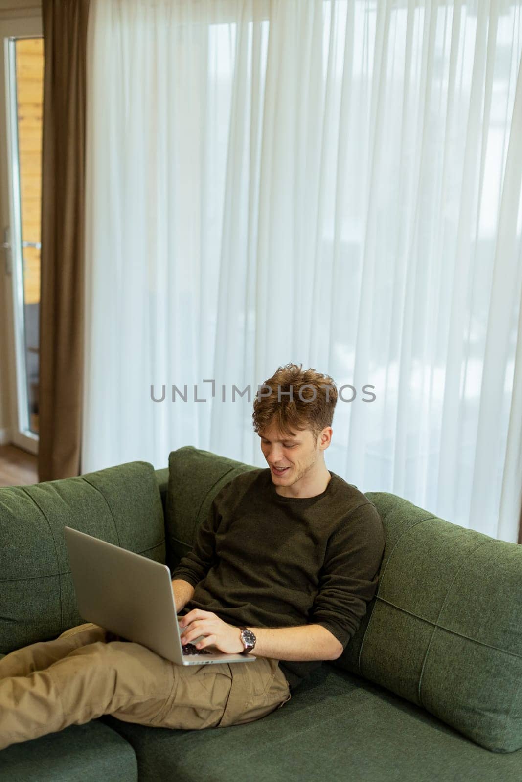 Focused on his laptop, a young man sits comfortably on a lush green couch, surrounded by light flooding through sheer curtains in a modern living space